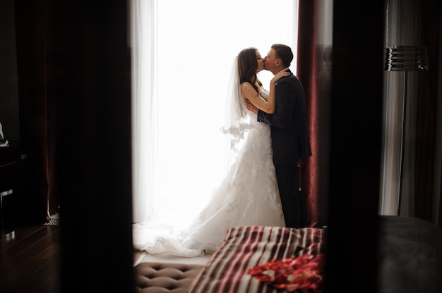 Bridegroom and bride in long dress are kissing near the window