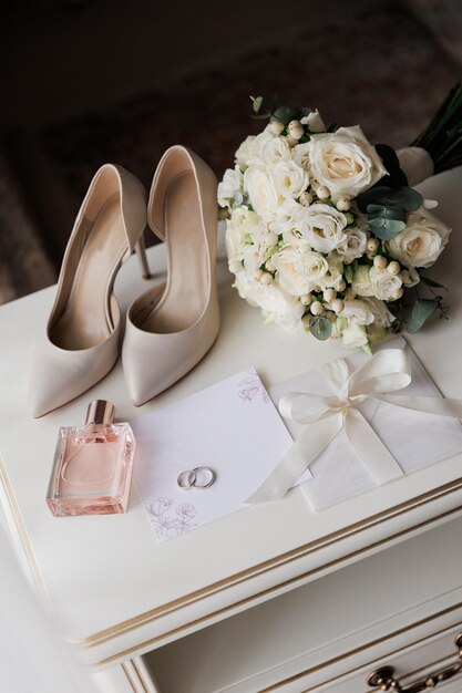 Photo brides wedding essentials heels bouquet rings perfume and invitation on a white table