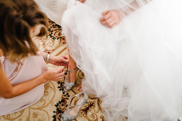 Bridesmaids put on leg bride stylish classic silver shoes Elegant Female putting hands on shoes on a background white dress Wedding morning preparation