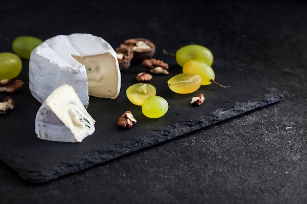 Brie type of cheese. Soft cheese with grapes and walnuts on black background. Copy space