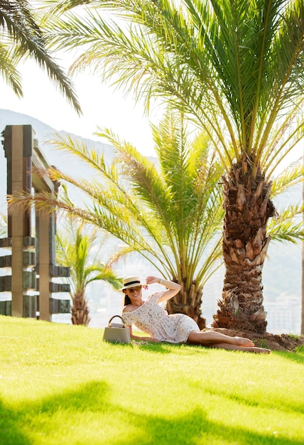 A bright beautiful girl in a light dress and hat lies on the grass under a palm tree in monaco in su