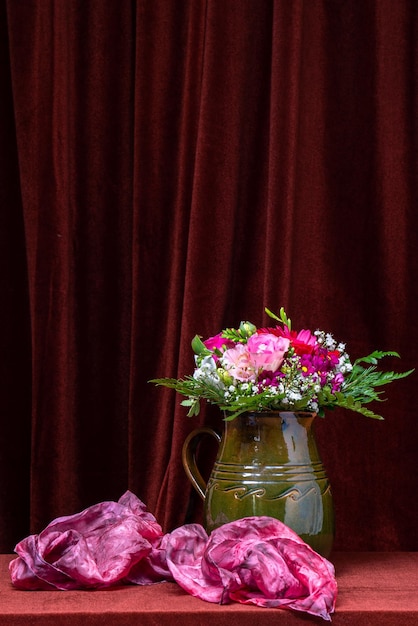 A bright bouquet of flowers in a green vase against a dark fabric background next to a pink scarf