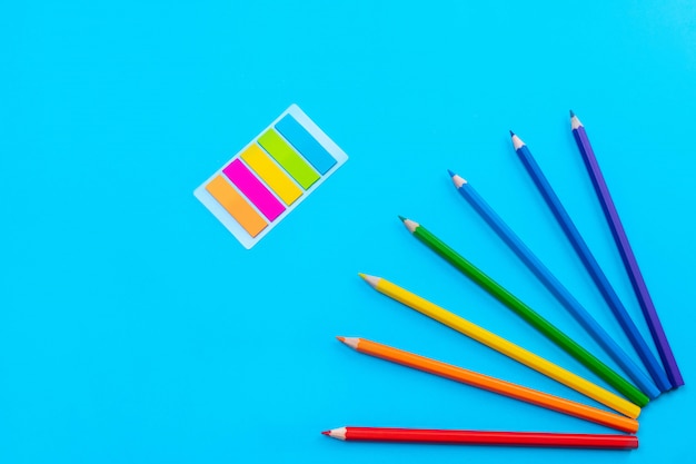 Bright colored pencils are laid out in the form of the sun in the right corner on a blue wall in the center of which are colored stickers in the form of stripes