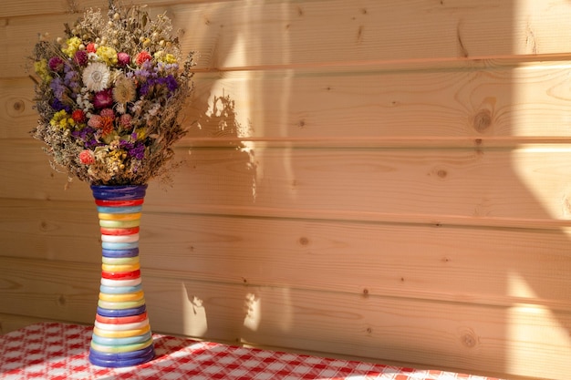 Bright flowers in colorful vase against wooden panel wall on red and white checked tablecloth.