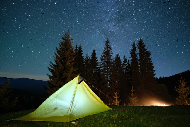 Bright illuminated tourist tent near glowing bonfire on camping site in dark mountains under night sky with sparkling stars. Active lifestyle concept.