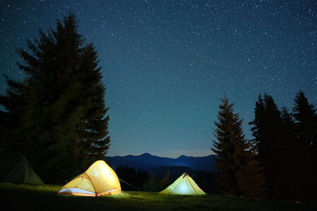 Bright illuminated tourist tents glowing on camping site in dark mountains under night sky with sparkling stars. Active lifestyle concept.