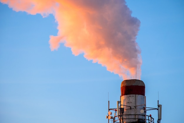 Bright orange smoke and steam from a high chimney of a power plant against a bright blue sky