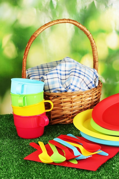 Bright plastic disposable tableware and picnic basket on the lawn on colorful background