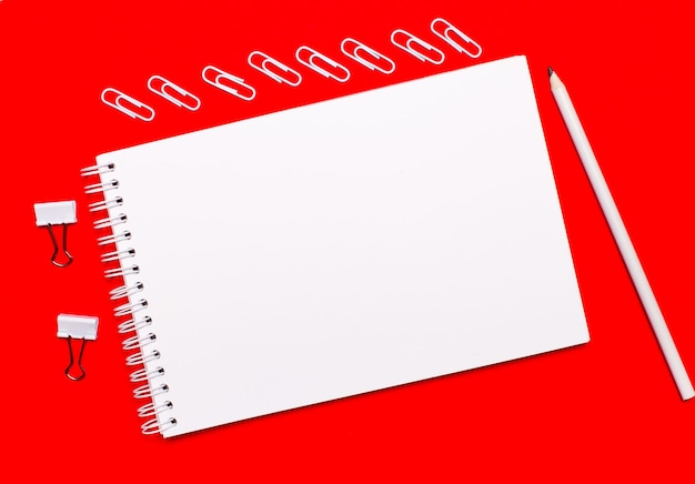 On a bright red background, a white pencil, white paper clips, white paper clips, and a white blank notebook with space to insert text.