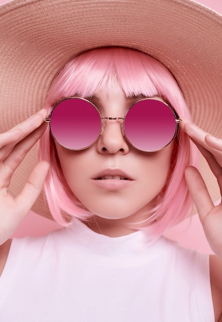 Bright summer portrait of a positive, gorgeous girl with pink hair, sunglasses and a braided hat on studio colorful