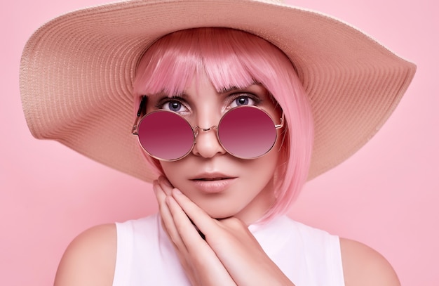 Bright summer portrait of a positive, gorgeous girl with pink hair, sunglasses and a braided hat on studio colorful