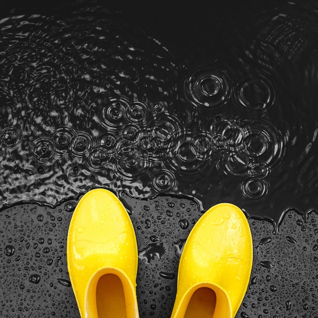 Bright yellow rubber boots stand in the rain on a black background covered with drops