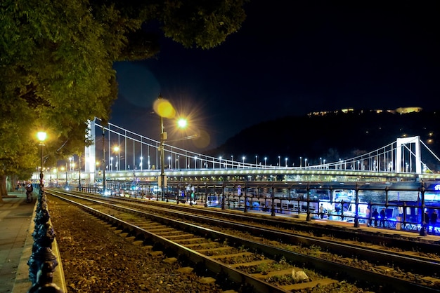 A brightly lit night bridge near a dark mountain In the foreground are the tram rails in perspective