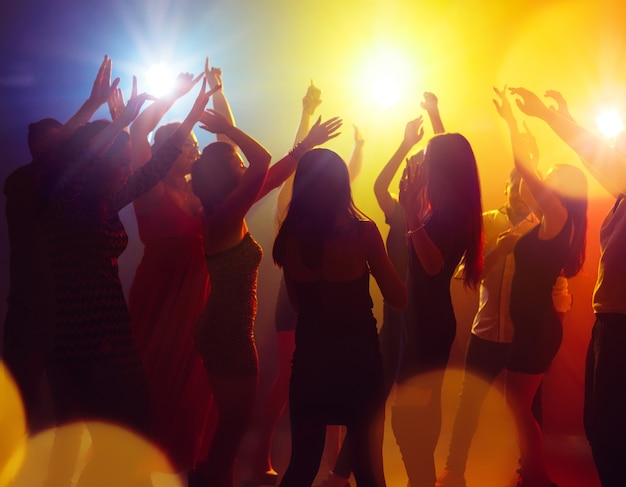 Brightness. A crowd of people in silhouette raises their hands on dancefloor on neon light background. Night life, club, music, dance, motion, youth. Yellow-blue colors and moving girls and boys.