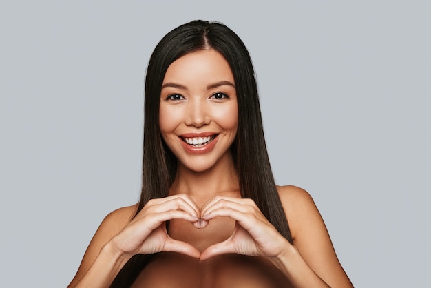 Bringing love. Beautiful young Asian woman gesturing and smiling while standing against grey background