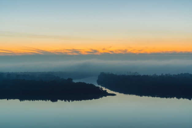 Broad river flows along shore with forest under thick fog. Early blue sky reflected in water. Yellow glow in picturesque predawn sky. Colorful morning mystical atmospheric landscape of majestic nature