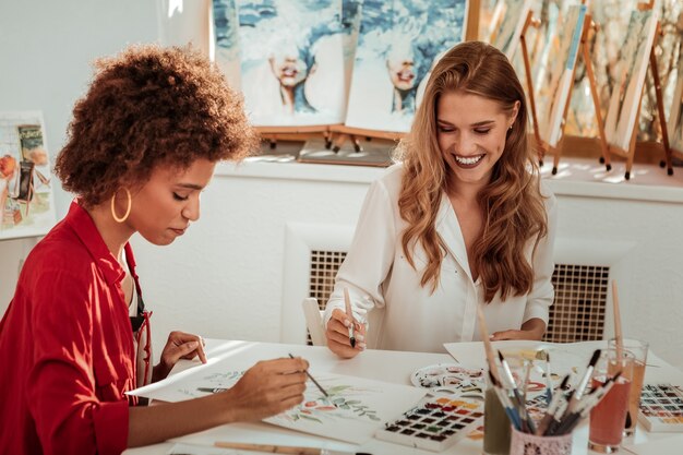 Broad smile. Beaming blonde-haired artist wearing white blouse smiling broadly painting with friend