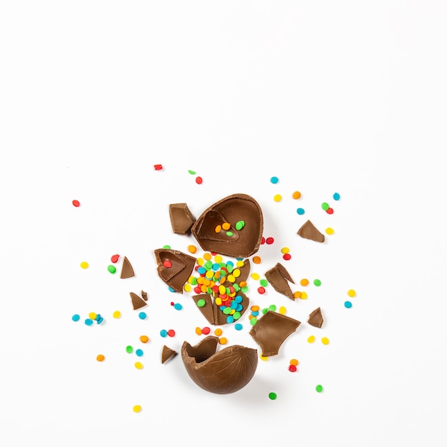 Photo broken easter chocolate egg and colorful decorations on a light surface. easter concept, easter treats. square. flat lay, top view