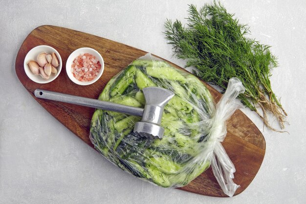 Broken salted cucumbers in a plastic bag on a wooden cutting board with a kitchen chop hammer surrounded by ingredients garlic pink salt dill