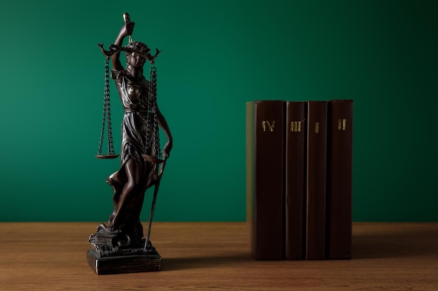 Bronze statuette with scales of justice and volumes of brown books on wooden table on dark green