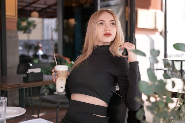 Brooding attractive young blonde woman sits at table in coffee shop with cup of cappuccino coffee latte