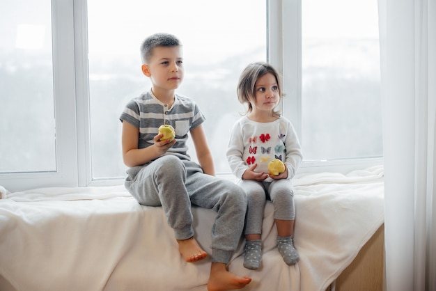 Brother and sister are sitting on the windowsill playing and eating apples. Happiness