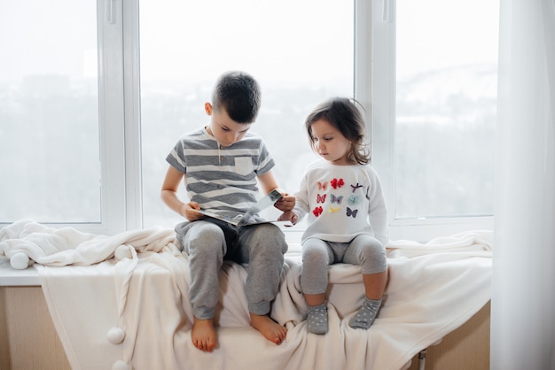 Brother and sister are sitting on the windowsill and reading a book. Happiness, family