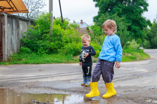 Brother and sister in rubber boots and raincoat are playing in a puddle