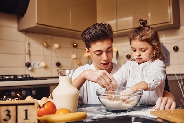 Brother with his little sister preparing food by using flour on kitchen and have fun.