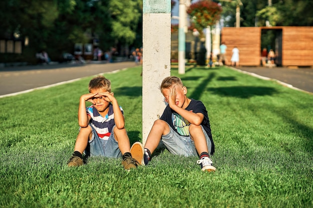 Brothers sit on park green lawn in public park and close eyes against sunlight