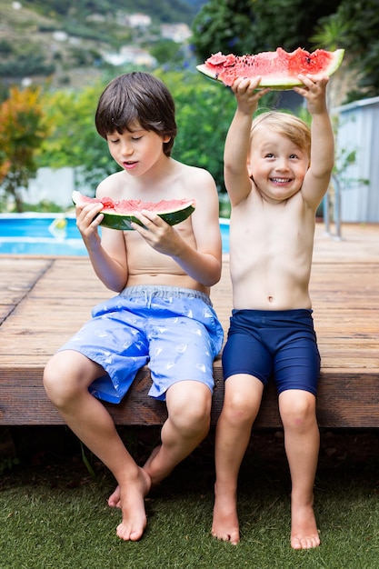 Brothers with big slices of watermelon sitting near the swimming pool healthy food concept siblings have a snack together outdoor in summer holidays