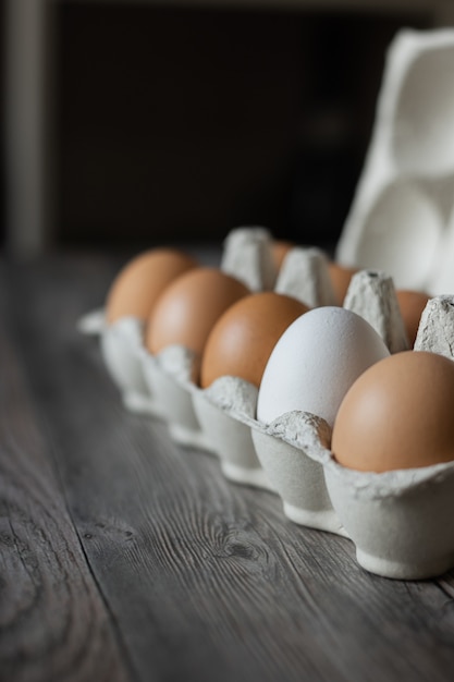 Brown chicken eggs and one white egg in a carton on wooden surface.