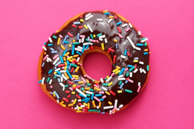 Brown chocolate donut with colorful sprinkles on a pink background.