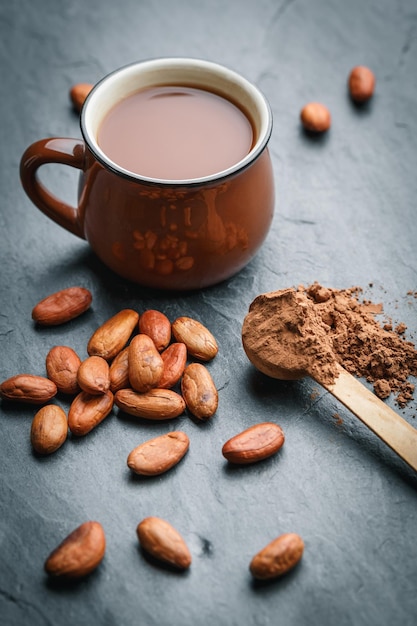 Brown cup of fresh cocoa drink with cocoa beans and powder in small wooden spoon on black stone background