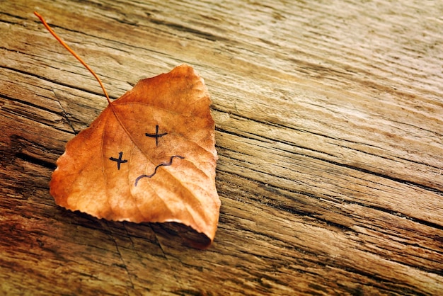 Brown dried leaf with a picture of a sad face on the old wooden background with cracks