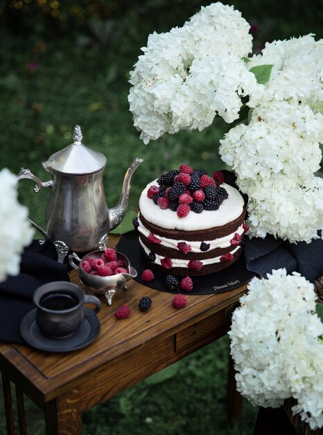 Brown fruit cake on the wooden narrow table in the garden. Silver tea pot and two black cups of tea are complementing the composition. 