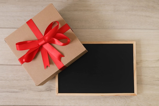 Brown gift box with red ribbon and blackboard on wooden background.