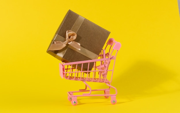 Brown gift box with a silk bow in a miniature metal trolley on a yellow background. Party Backdrop, surprise, seasonal sale