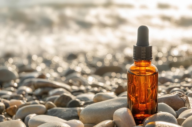 Brown glass cosmetic bottle with black pipette on stone pebble beach on sunny day. Natural beauty concept.