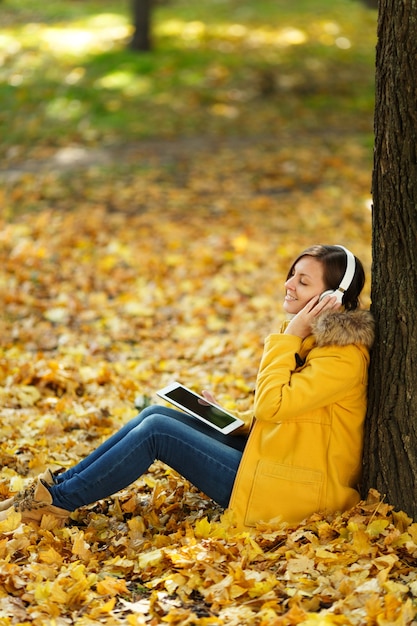 Photo the brown-haired smiling woman in yellow coat and jeans sitting and listening to music under a tree with a tablet in her hands and headphones in fall city park on a warm day. autumn golden leaves.