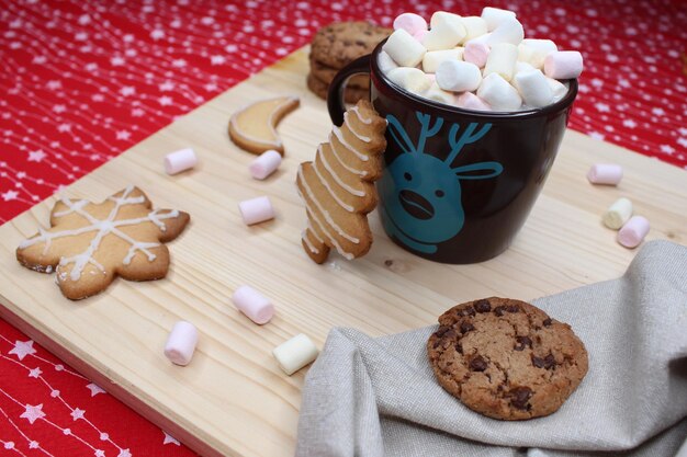 Brown mug with marshmallows Cookies with Christmas shapes Gray kitchen rag Wood and red tablecloth