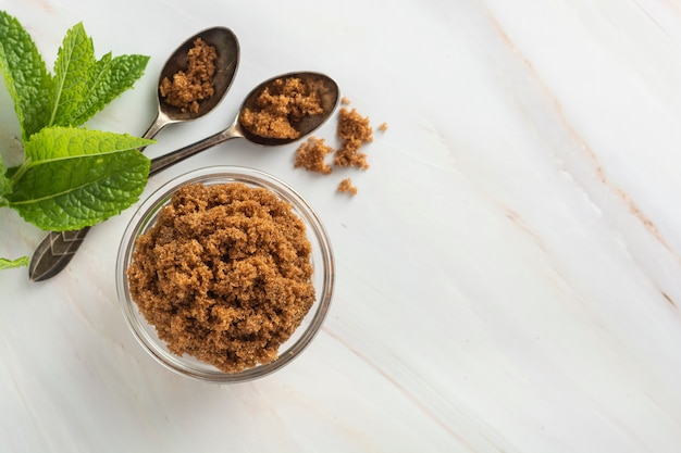 Brown sugar isolated in glass bowl, over styled marble background. Mint leaves. Top view, copy space.