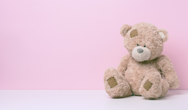 Brown teddy bear with patches sits on a white table, pink background, copy space