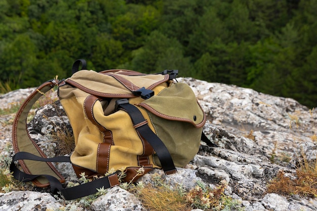 A browngreen backpack lies on a large gray stone on a rock against the backdrop of green mountains