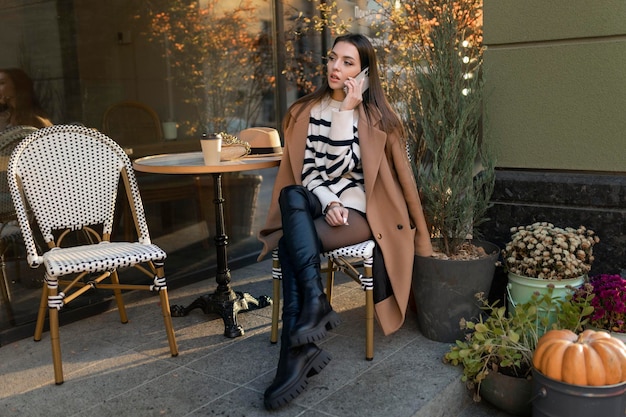 A brunette beautiful stylish caucasian woman in casual clothes on a walk along the street of a European city sits in a cafe and talks on the phone