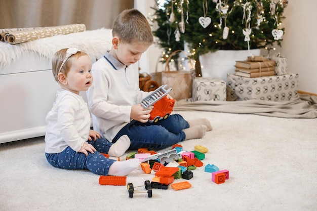 Brunette boy and girl sitting near christmas tree at home. Siblings are playing together. Boy and girl wearing blue and beige clothes.