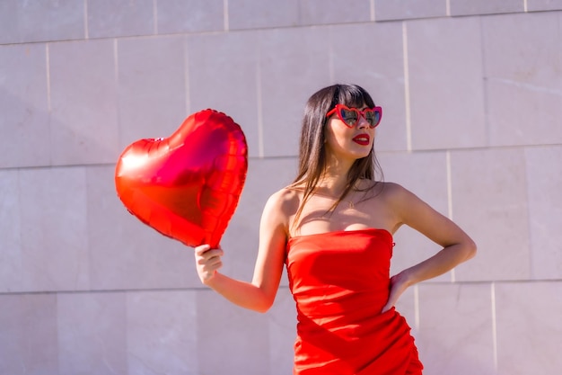 Brunette caucasian girl with a heart balloon on Valentines day sunglasses and red dress Lifestyle of people in love on a gray background