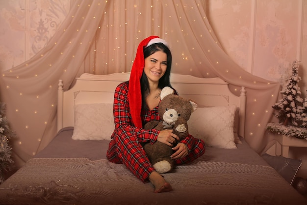 brunette in Christmas pajamas Santa Claus hat sitting on the bed hugging a teddy bear