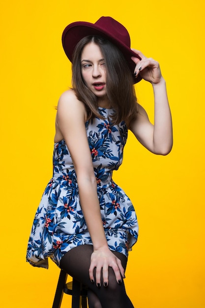 A brunette in a dress and hat is sitting on a chair on a yellow backgroundFashion shooting of a young woman