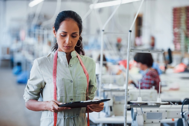 Brunette dressmaker works in the factory and holds notepad in hands.